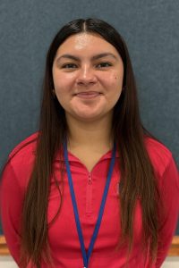 Portrait of smiling woman wearing pink top and blue lanyard in front of a blue wall.