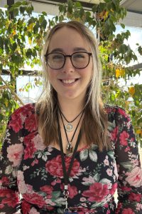 Portrait of a smiling woman with glasses in a floral dress, wearing a lanyard by a sunny window and leafy indoor plant.